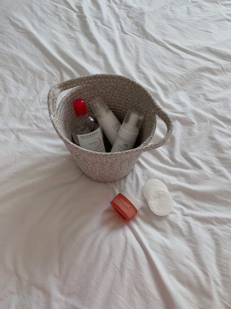 Skincare bottles arranged in a woven basket on white bed linen