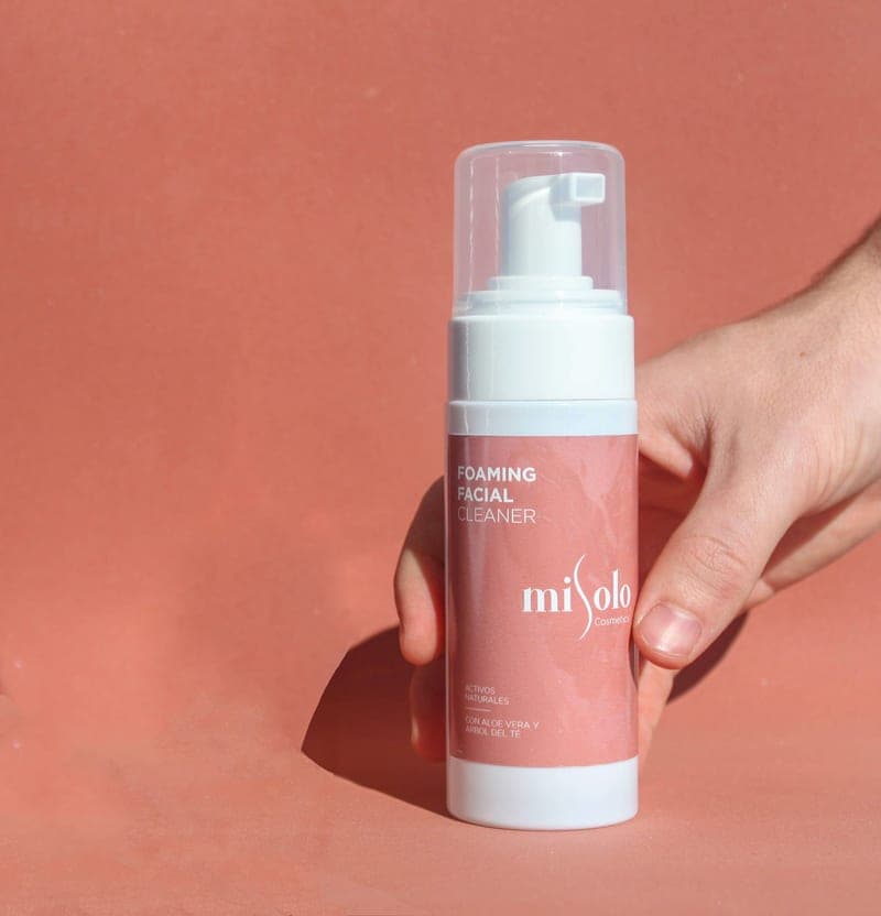 A hand holding a foaming facial cleanser bottle against a pale background