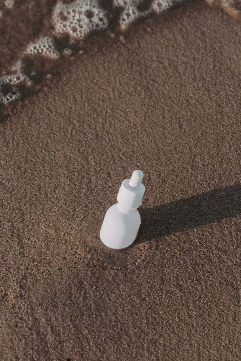 A plain white cosmetic bottle resting in sand near the sea